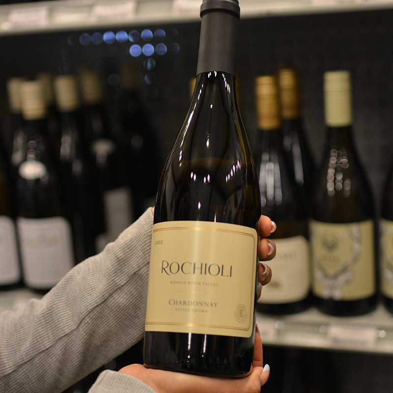 Hand holding a bottle of Chardonnay wine in front of a wine selection at Hickey's Wine & Spirits.